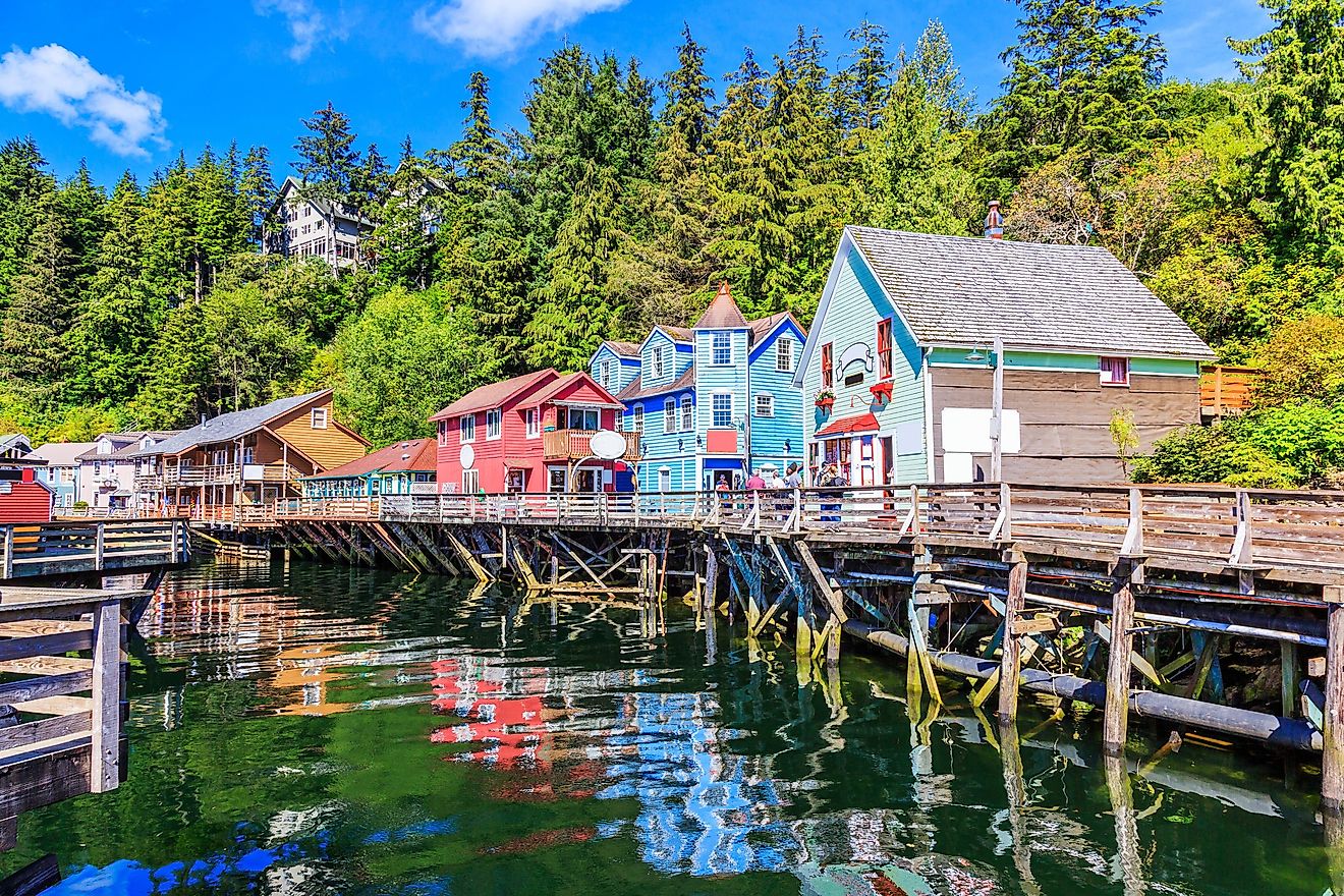 The waterside stores in Ketchikan, Alaska. Colorful wooden buildings on stilts line a scenic waterfront, reflecting in the clear water. Lush green forest and a blue sky complete the vibrant, tranquil scene.