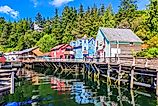 The waterside stores in Ketchikan, Alaska. Colorful wooden buildings on stilts line a scenic waterfront, reflecting in the clear water. Lush green forest and a blue sky complete the vibrant, tranquil scene.