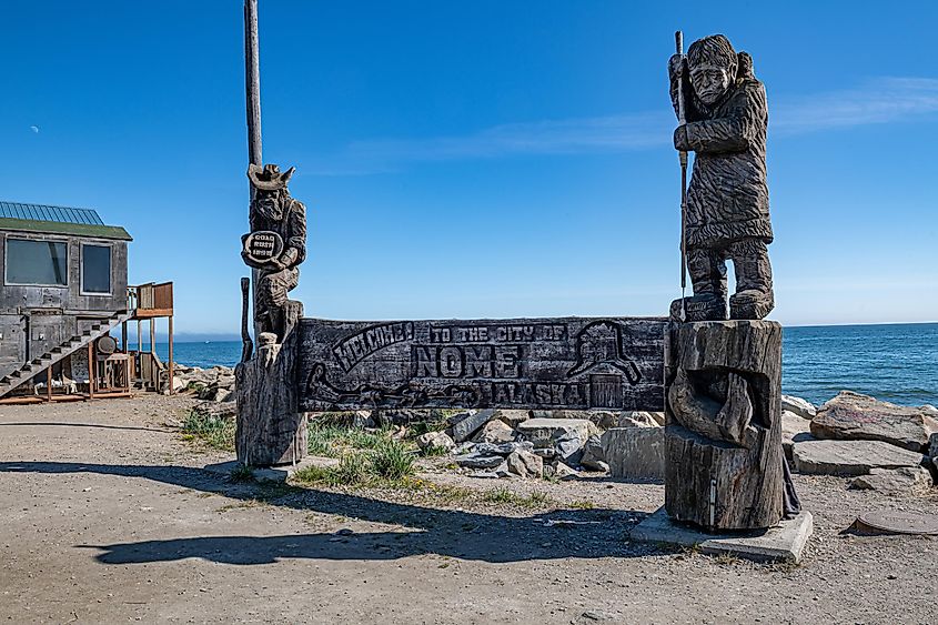 A wooden sign with carved figures welcomes visitors to Nome, Alaska, set against a clear blue sky and ocean backdrop, evoking a rustic, coastal charm.