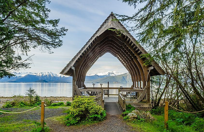 Wooden arch structure frames a serene view of a lake with snow-capped mountains. Surrounded by lush greenery, the scene feels peaceful and inviting.