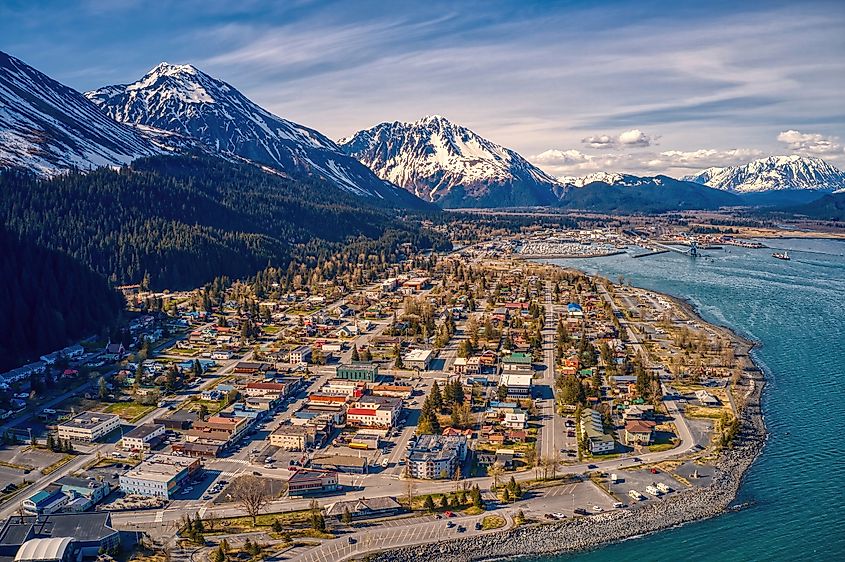Aerial view of a coastal town with colorful buildings and streets, surrounded by snow-capped mountains and a blue bay. The scene is serene and picturesque.
