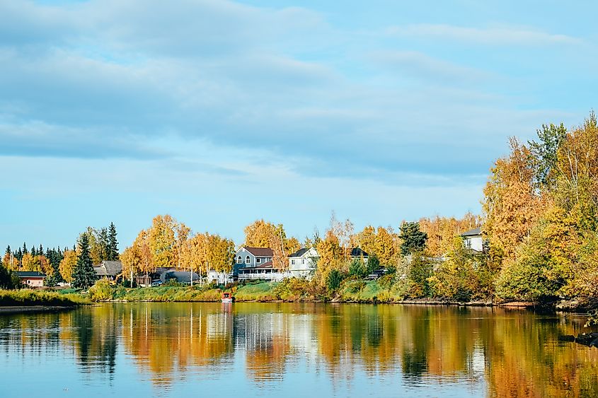 Tranquil autumn scene with colorful trees, houses, and their reflections on a calm lake under a partly cloudy blue sky. The mood is peaceful and serene.