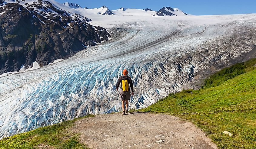 A person with a yellow backpack stands on a trail overlooking a vast glacier, surrounded by rocky mountains. The scene conveys a sense of awe and tranquility.